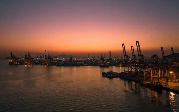 Container port illuminated at sunset, with cargo cranes silhouetted around a busy harbour.
