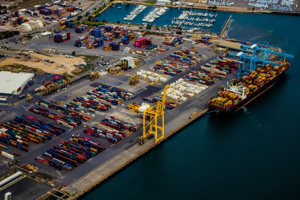 Aerial view of a container terminal with stacked shipping containers, cargo cranes, and a docked container ship beside a marina.