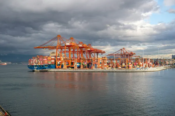 Container ship docked at a busy port terminal beneath large cargo cranes and dark storm clouds.