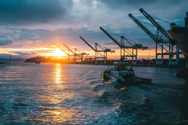 Tugboat moving through a busy container port at sunset, with cargo cranes lining the waterfront.