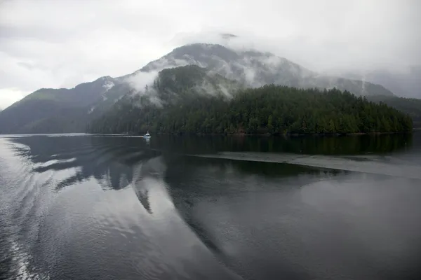 Mist-covered, forested mountains reflected in calm coastal waters as a small boat travels through the inlet.