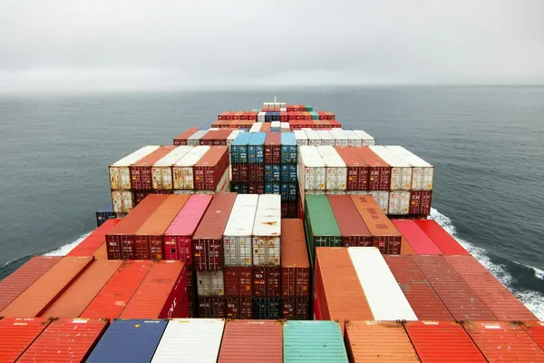 View across stacked shipping containers aboard a cargo vessel travelling through open ocean.