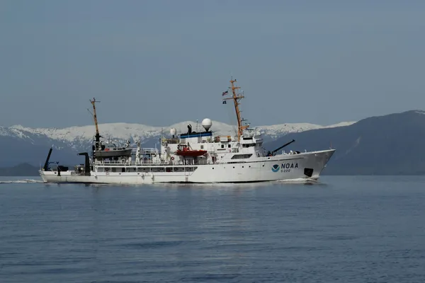 NOAA oceanographic research vessel underway in calm coastal waters, with snow-covered mountains in the background.