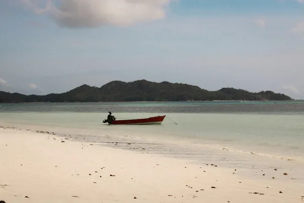 Small red motorboat anchored in shallow turquoise water beside a sandy beach, with a forested island in the distance.
