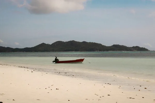 Small red motorboat anchored in shallow turquoise water beside a sandy beach, with a forested island in the distance.