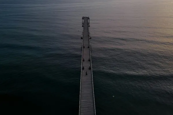 Long wooden pier extending into calm ocean water at dusk, viewed from above.