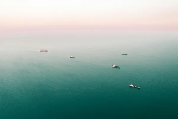 Aerial view of several cargo ships spread across calm open water beneath a hazy sky.