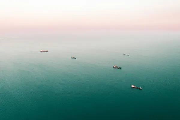 Aerial view of several cargo ships spread across calm open water beneath a hazy sky.